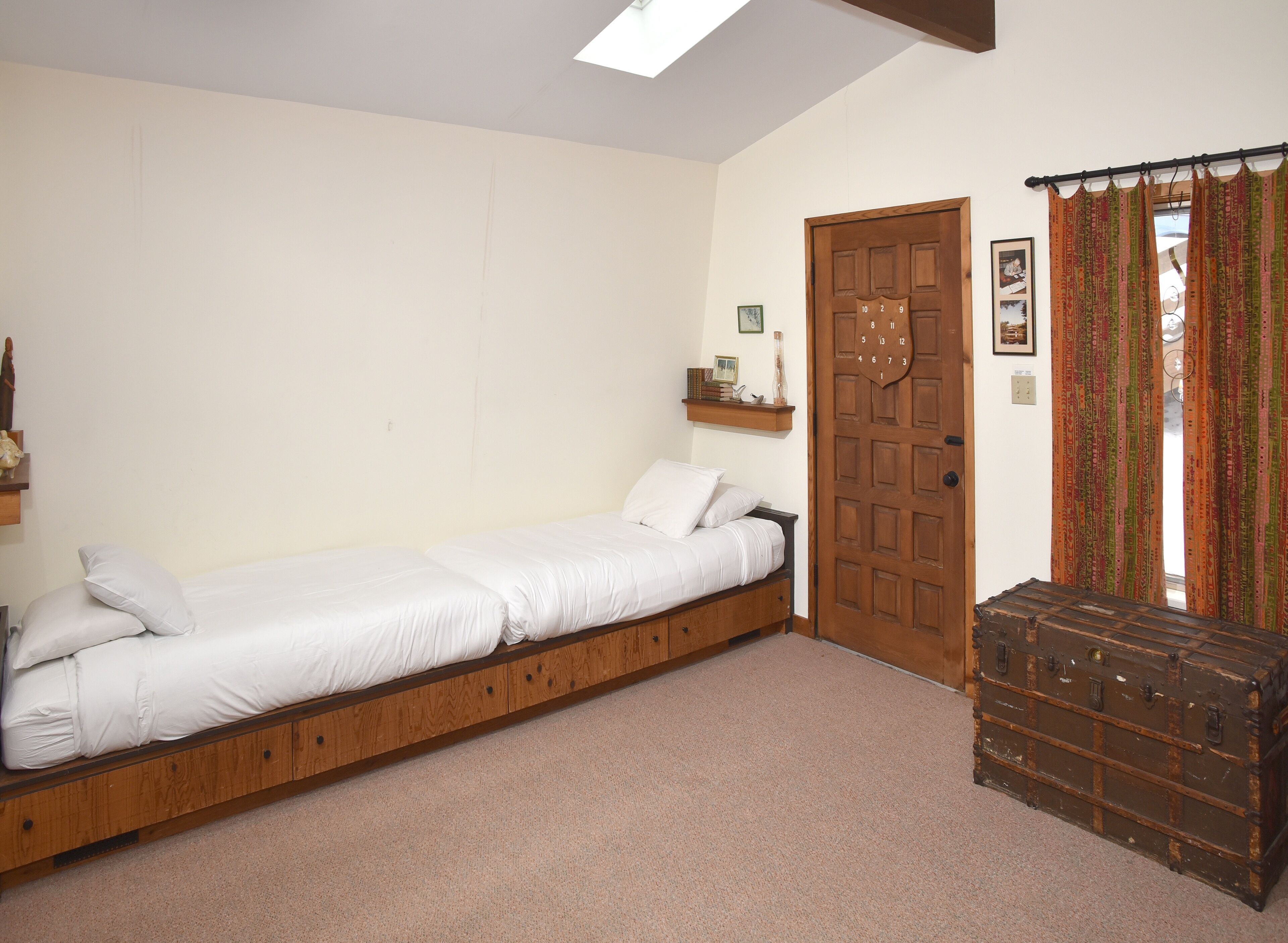 A single bed with a wooden frame and a headboard, a wooden chest, a door with a clock, and a shelf with books and a vase.