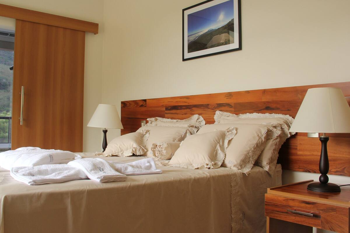 Una cama bien hecha con sábanas blancas y almohadones decorativos, un cabecero de madera, dos lámparas de noche y un cuadro en la pared.