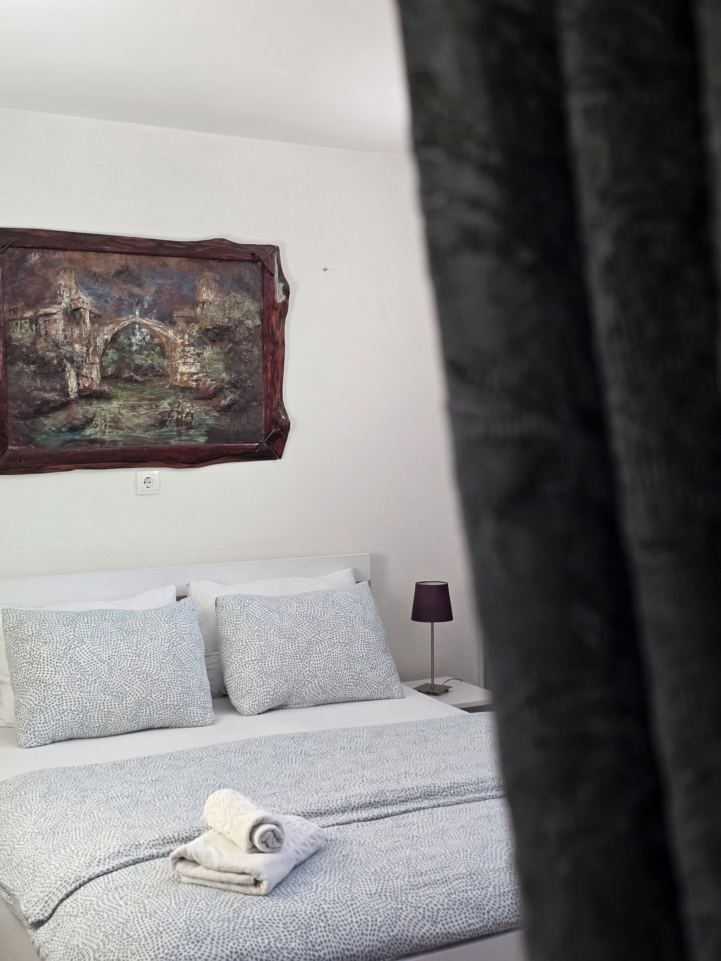 A neatly made bed with patterned pillows, a painting of a bridge on the wall, and a lamp on the nightstand.