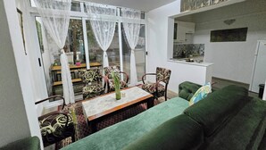 A living room with a green sofa, a dining table with chairs, and a view through large windows.