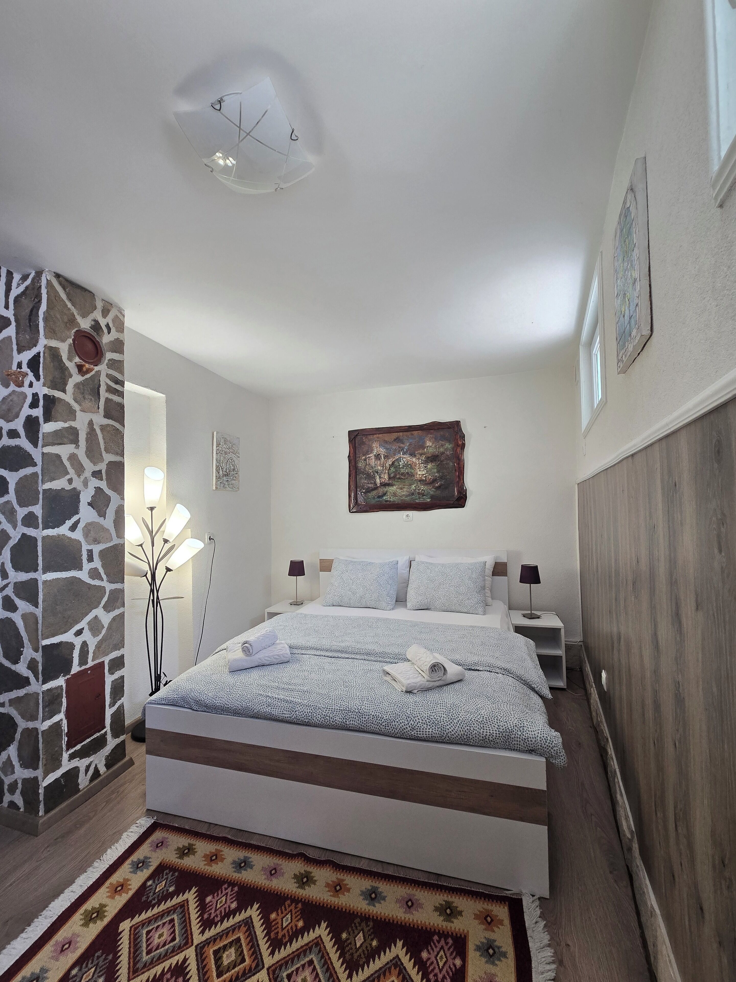A bedroom with a stone accent wall, a bed with patterned bedding, a nightstand with lamps, and a painting on the wall.