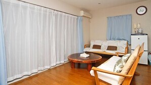Una habitación con piso de madera, una cama con cabecera blanca y marrón, una pequeña mesa redonda cubierta con un mantel blanco, un equipo de aire acondicionado blanco y un refrigerador blanco.