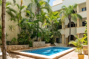 A pool area with a stone-lined edge, surrounded by lush tropical plants and a multi-story building with balconies. - Piedra Viva by Playa Paradise (Playa del Carmen)