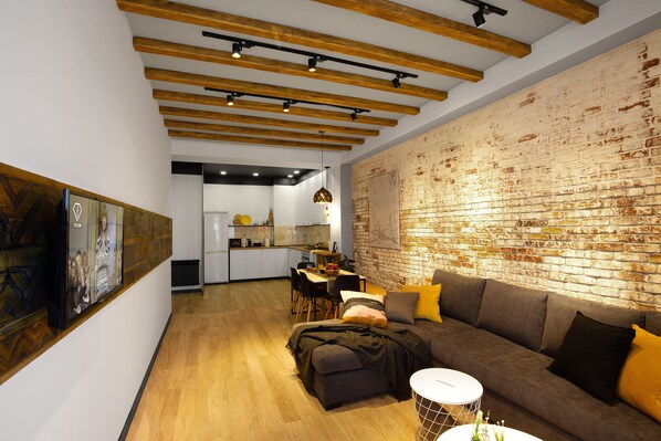 A modern living room with a brick wall, wooden beams, and a dining area with a kitchen in the background.