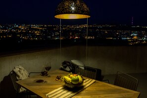 A dining table with a bowl of fruit and wine glasses, a lamp, and a view of a cityscape at night.