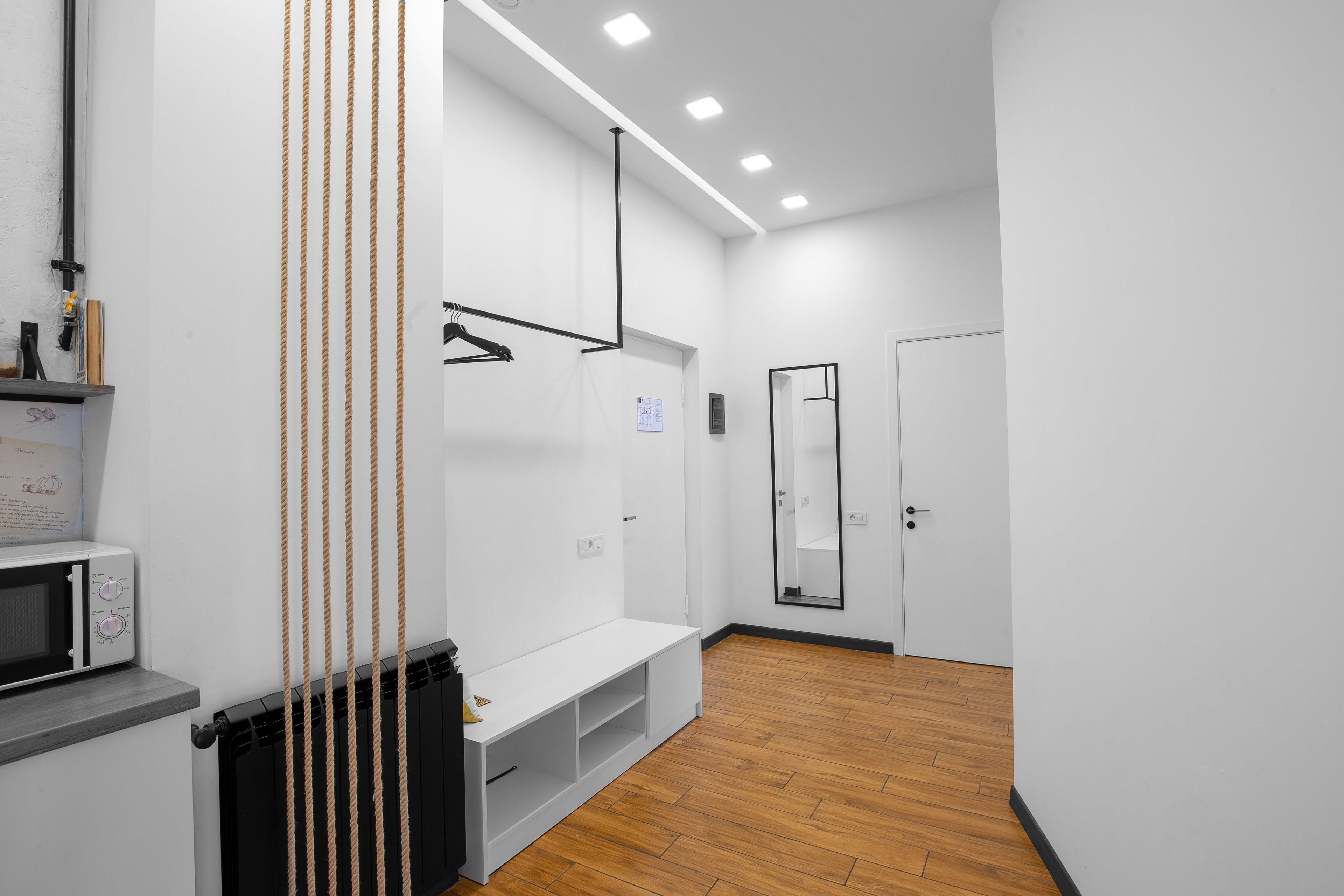 A modern, minimalist room with a white wall, a wooden floor, a white bench, a black radiator, and a white microwave.
