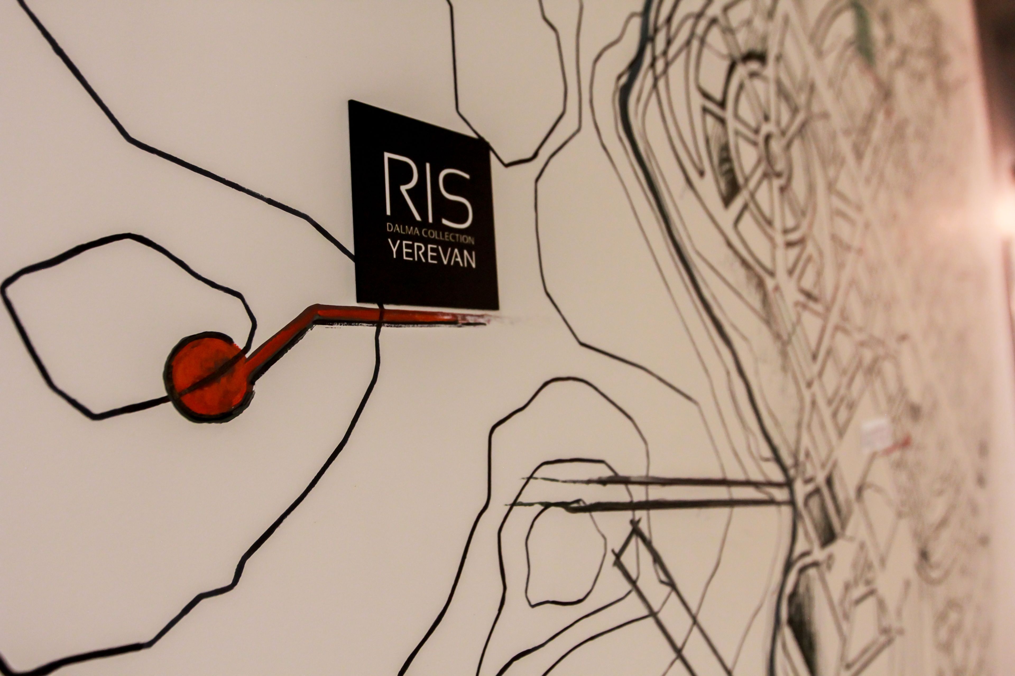 Una pared con un letrero negro que dice "RIS DALMA COLLECTION YEREVAN" y un objeto rojo parecido a una llave.