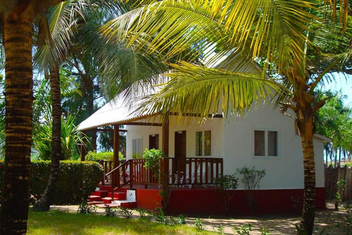 Una casa blanca con porche rojo y escalones, rodeada de palmeras y follaje.