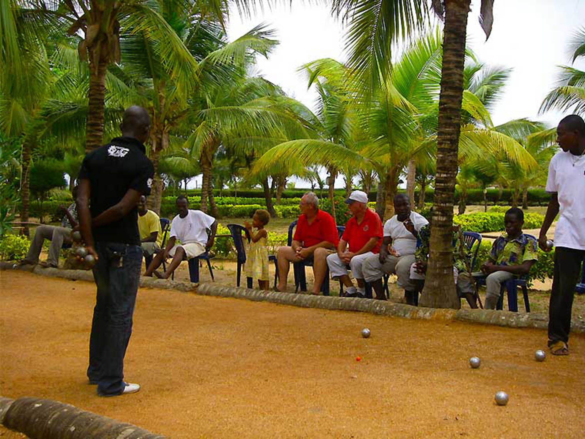 A group of people playing a game with metal balls on a sandy ground, surrounded by palm trees.