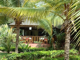 A small, single-story building with a wooden deck and a thatched roof, surrounded by lush tropical vegetation.