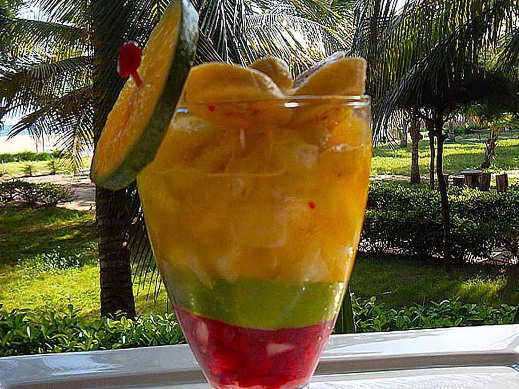 A layered fruit drink in a glass with a slice of lime on the rim, set against a background of palm trees and greenery.