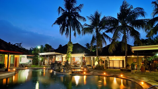 A pool area with palm trees, a building with a tiled roof, and a well-lit walkway. - Dewi Sinta Hotel and Restaurant by ABM (Tabanan)