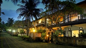 A nighttime view of a residential area with houses, palm trees, and a paved walkway. - Dewi Sinta Hotel and Restaurant by ABM (Tabanan)
