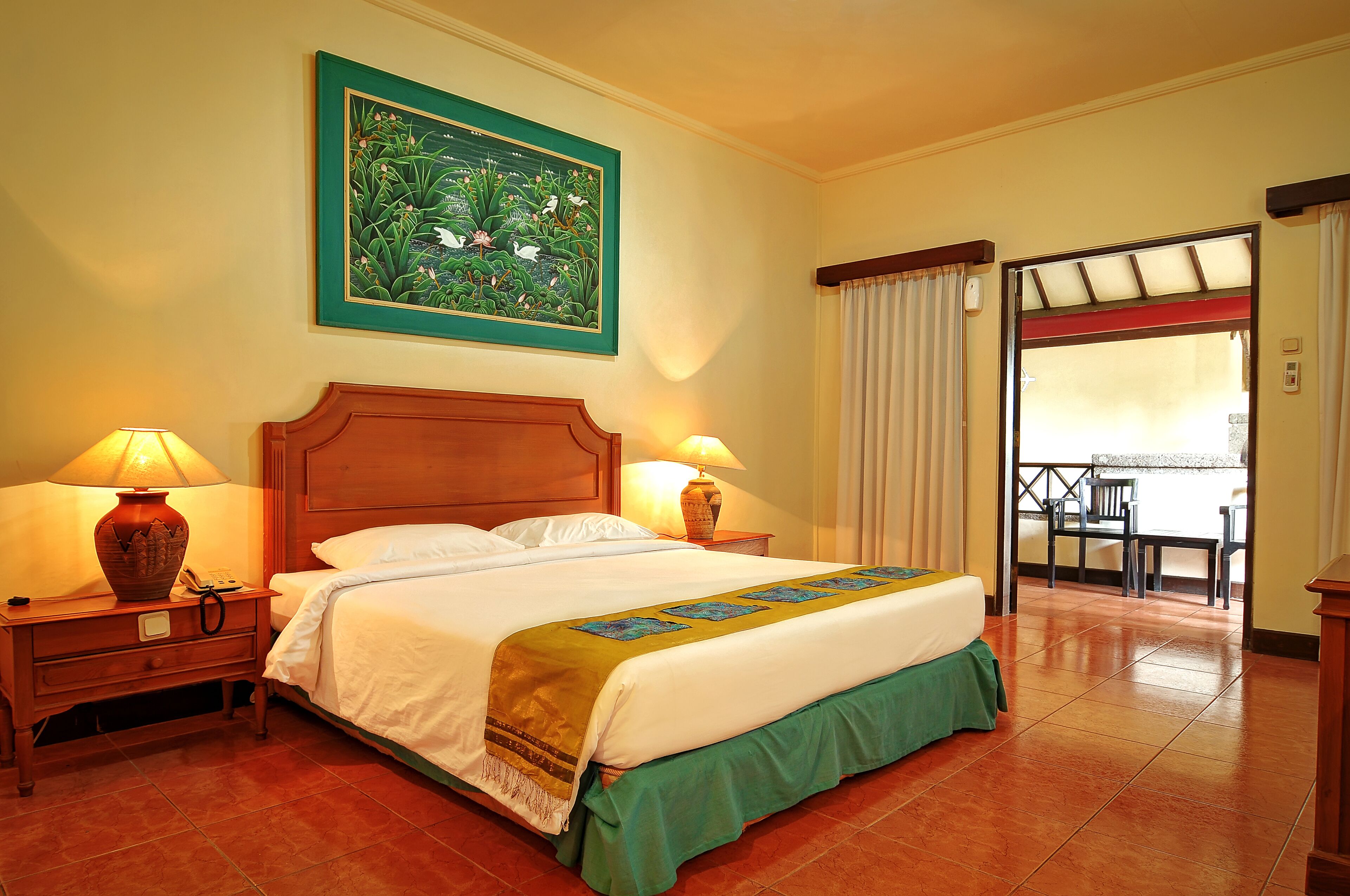 A hotel room with a bed, two bedside lamps, a nightstand, a green bed runner, a large floral painting on the wall, and a view of the outside balcony.