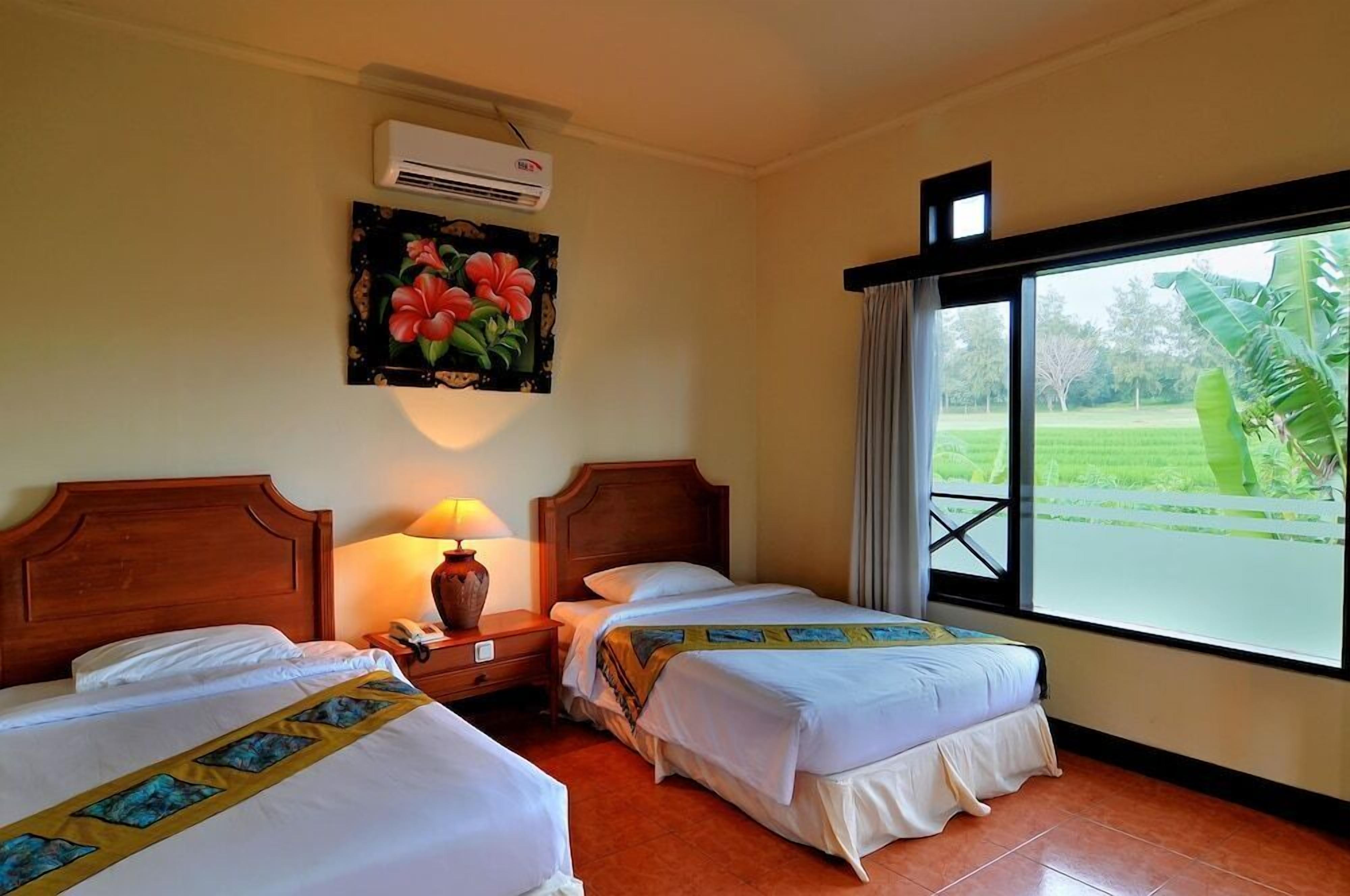 A hotel room with two beds, a nightstand with a lamp, an air conditioner, a painting of flowers, and a window with a view of greenery.