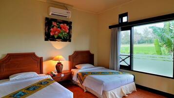 A hotel room with two beds, a nightstand with a lamp, an air conditioner, a painting of flowers, and a window with a view of greenery.