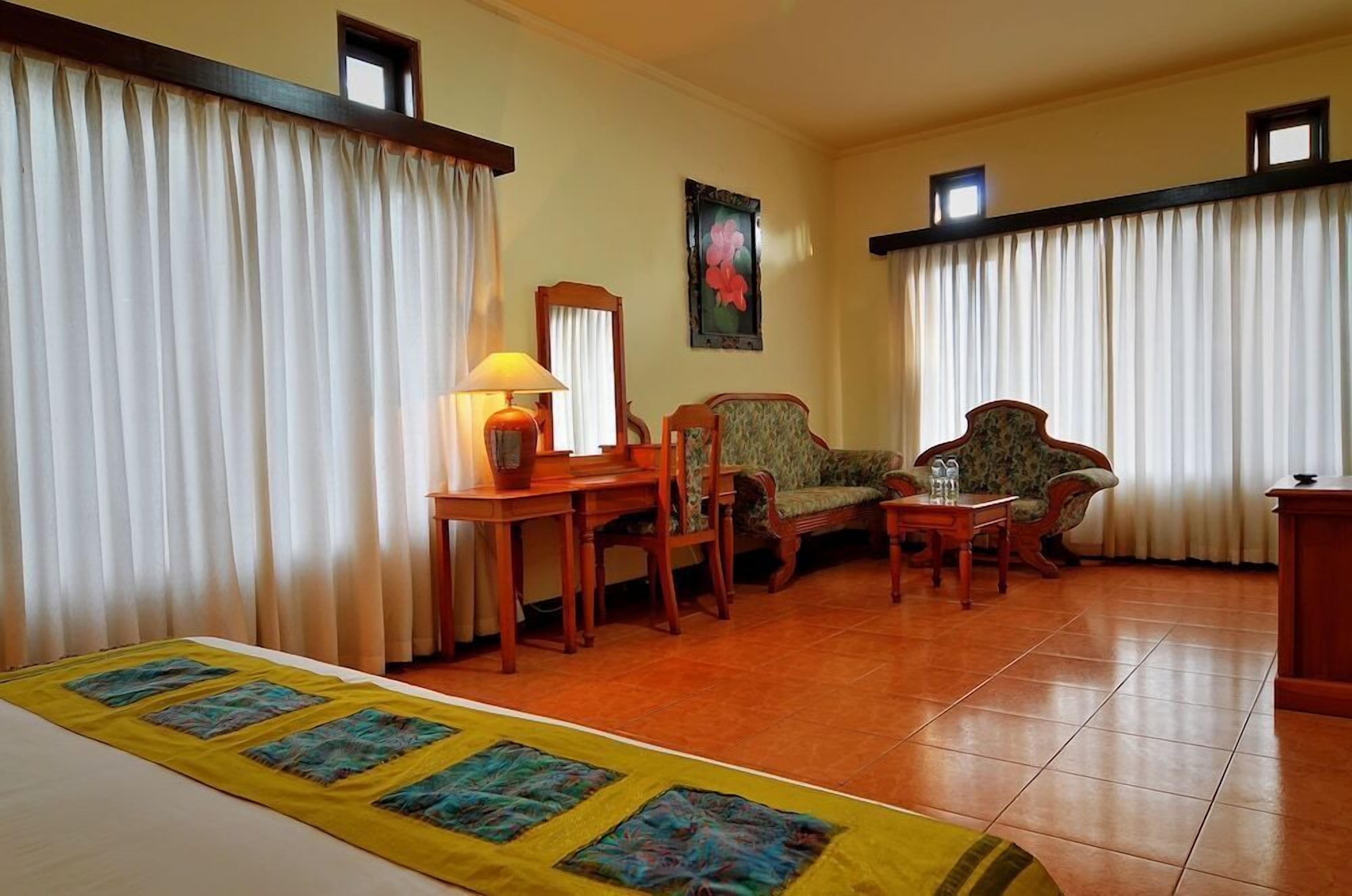 Une chambre d’hôtel avec un lit, un bureau avec une lampe, deux fauteuils, une petite table avec une bouteille et un tableau au mur.