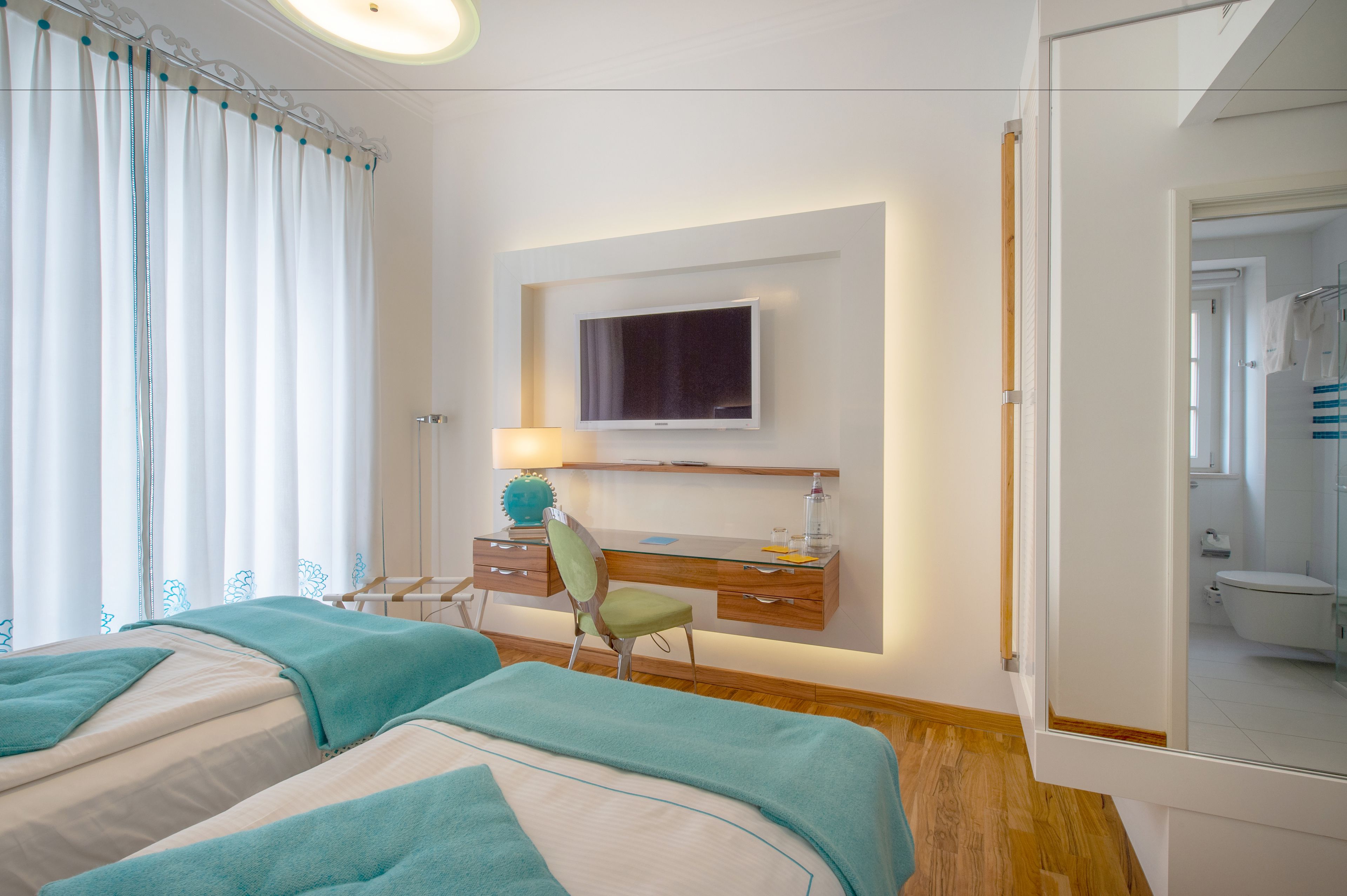 Hotellihuone, jossa on kaksi sänkyä, työpöytä, tuoli, televisio ja kylpyhuone, joka näkyy peilin kautta.