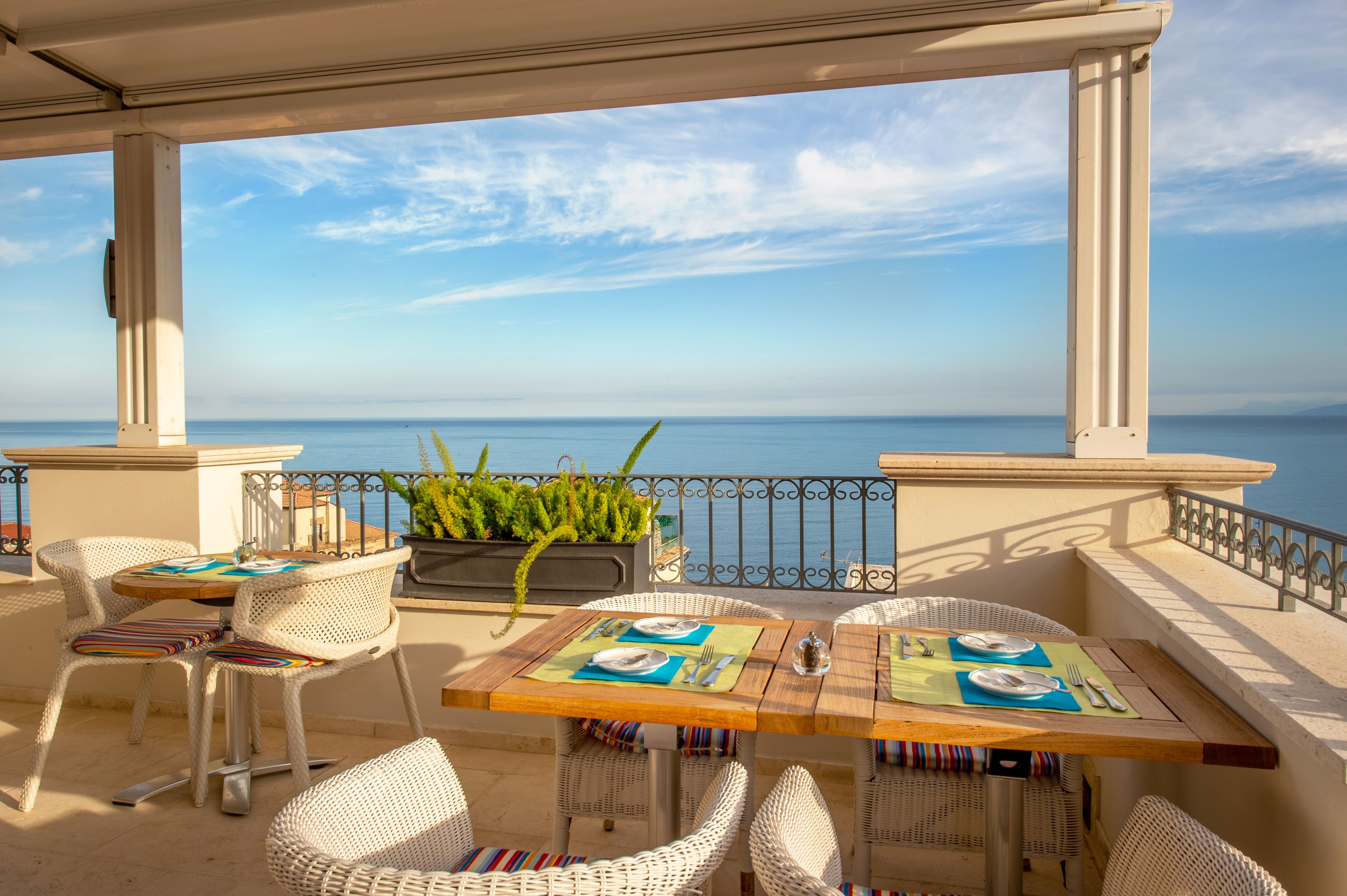 A terrace with wicker furniture, a wooden table set for dining, and a view of the sea.