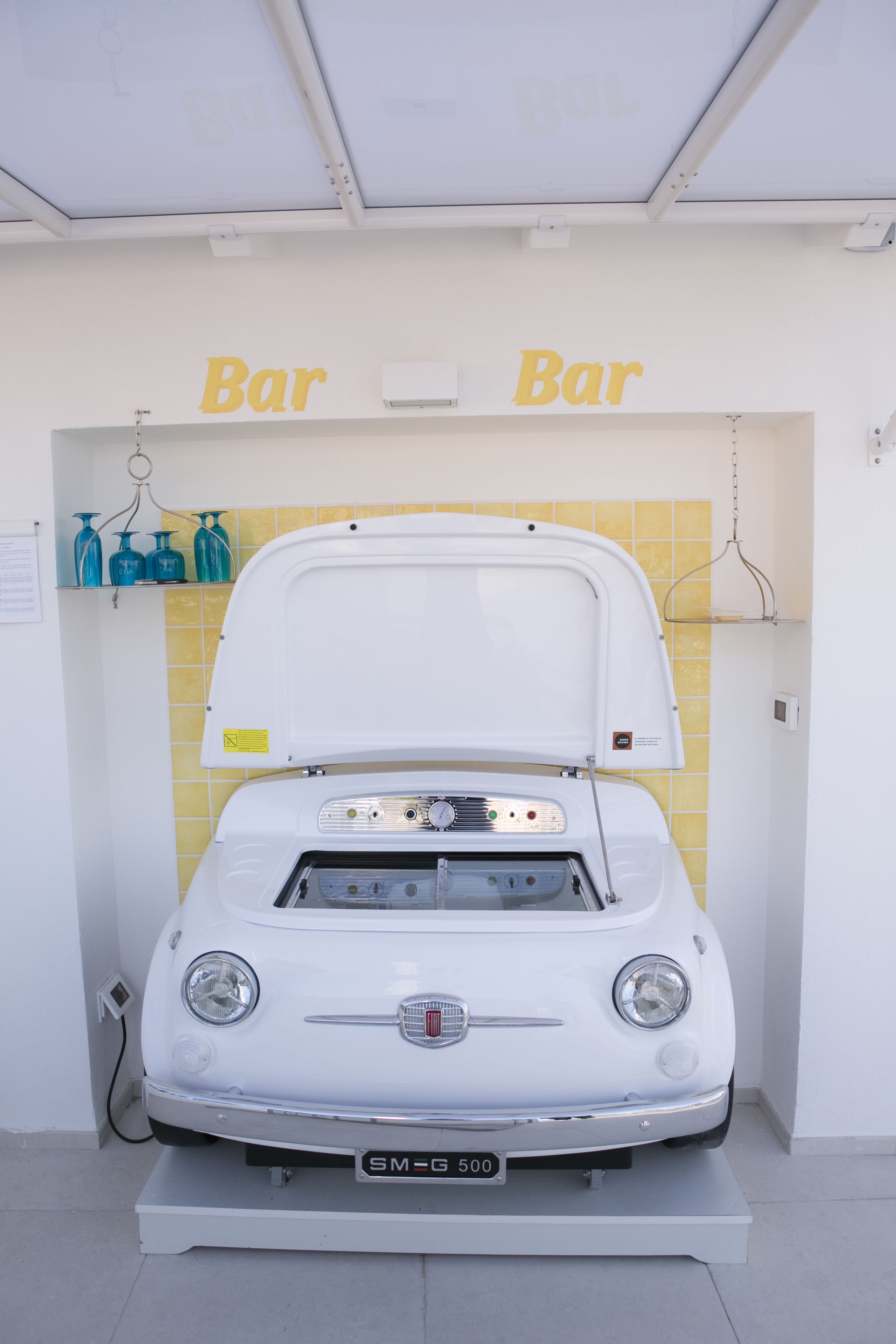 Een oude witte auto, motorkap open, staat in een kamer met gele tegelwanden en twee 'Bar'-borden erboven.