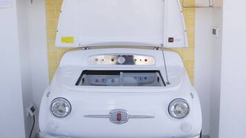 Een oude witte auto, motorkap open, staat in een kamer met gele tegelwanden en twee 'Bar'-borden erboven.