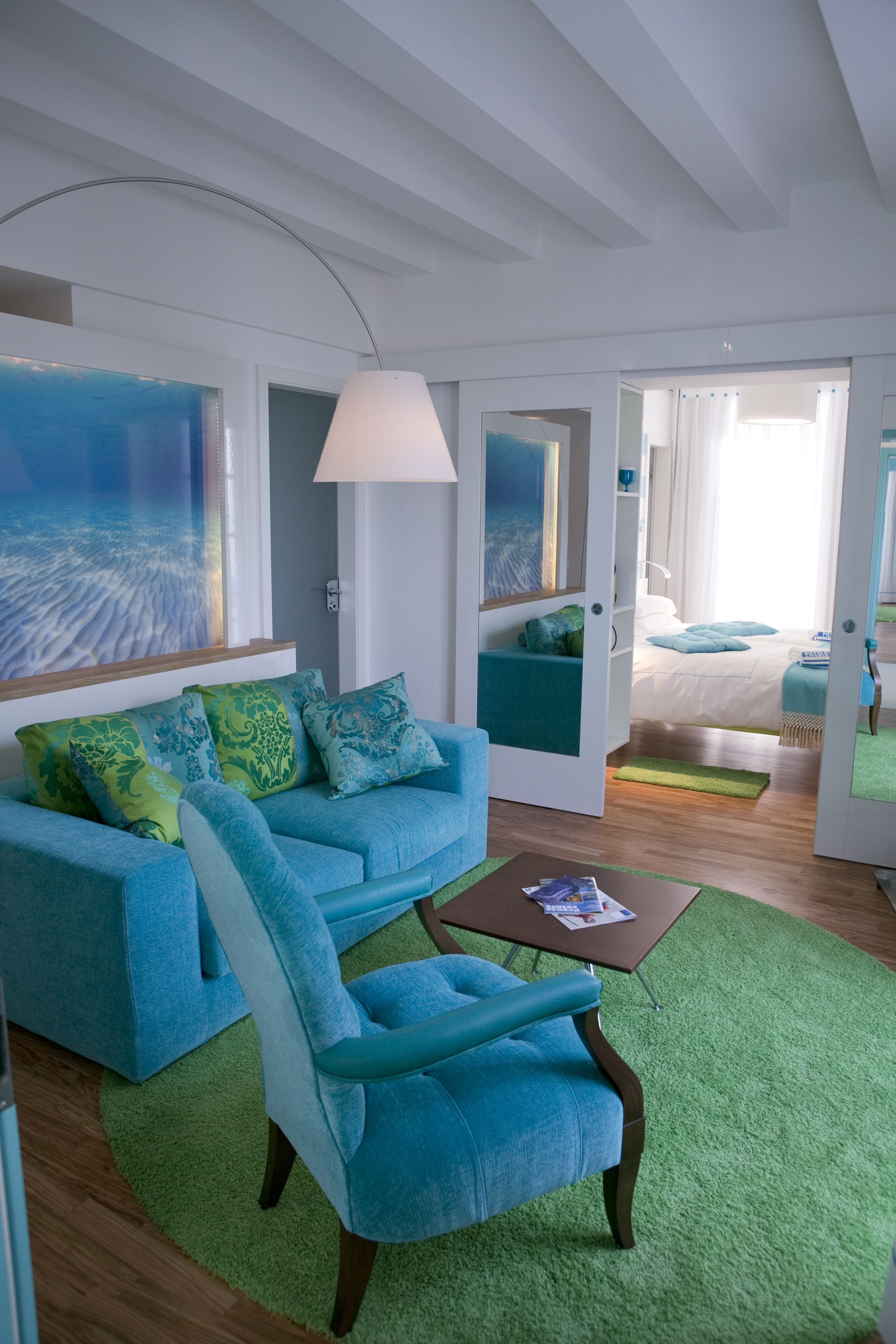 A modern living room with a blue sofa, a matching armchair, and a green rug. There is a painting on the wall and a lamp on a side table.