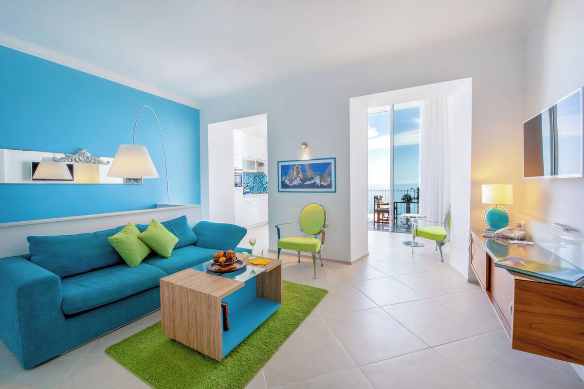 A modern living room with a blue sofa, green cushions, a wooden coffee table, and a green rug. There is a dining area with a table and chairs, and a balcony with a view.