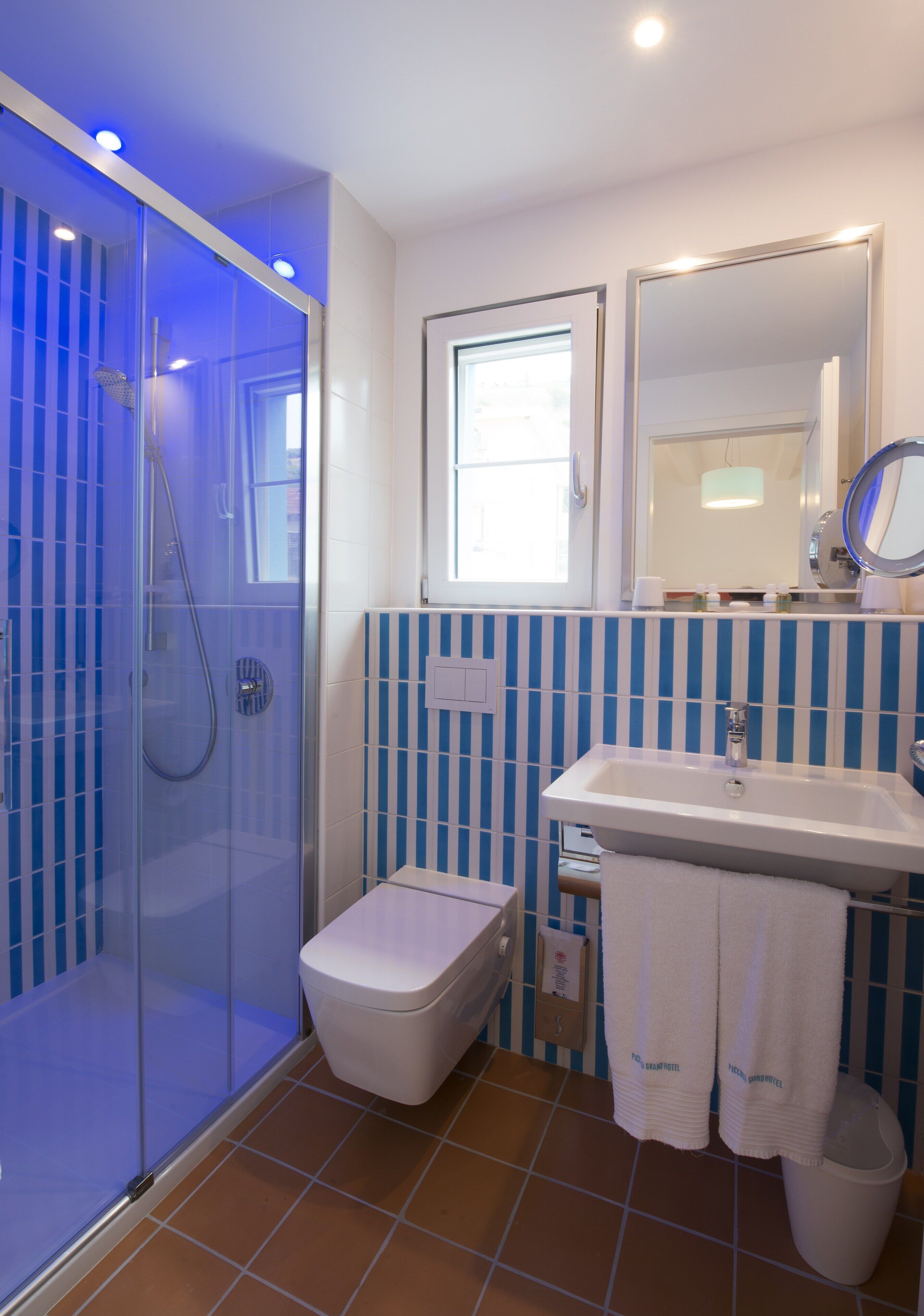 Un baño con ducha envuelta en vidrio, un inodoro blanco, un lavamanos con espejo y azulejos a rayas azul y blanco.