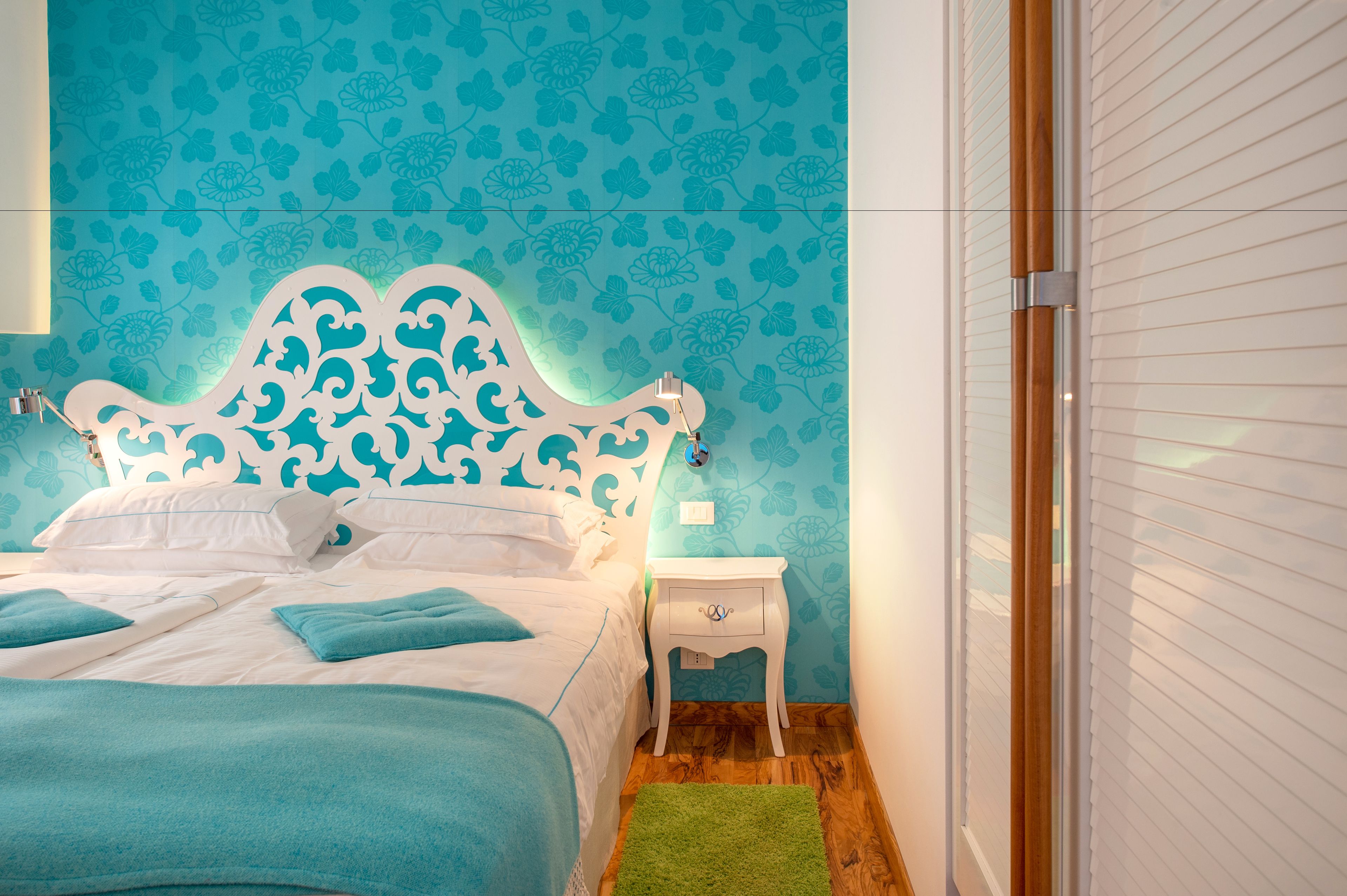 A neatly made bed with a white headboard and blue bedding, a small bedside table with a lamp, and a green rug on a wooden floor.