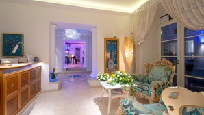 Un vestíbulo de hotel con una recepción, un área de estar con muebles ornamentados y una vista del área del bar al fondo.