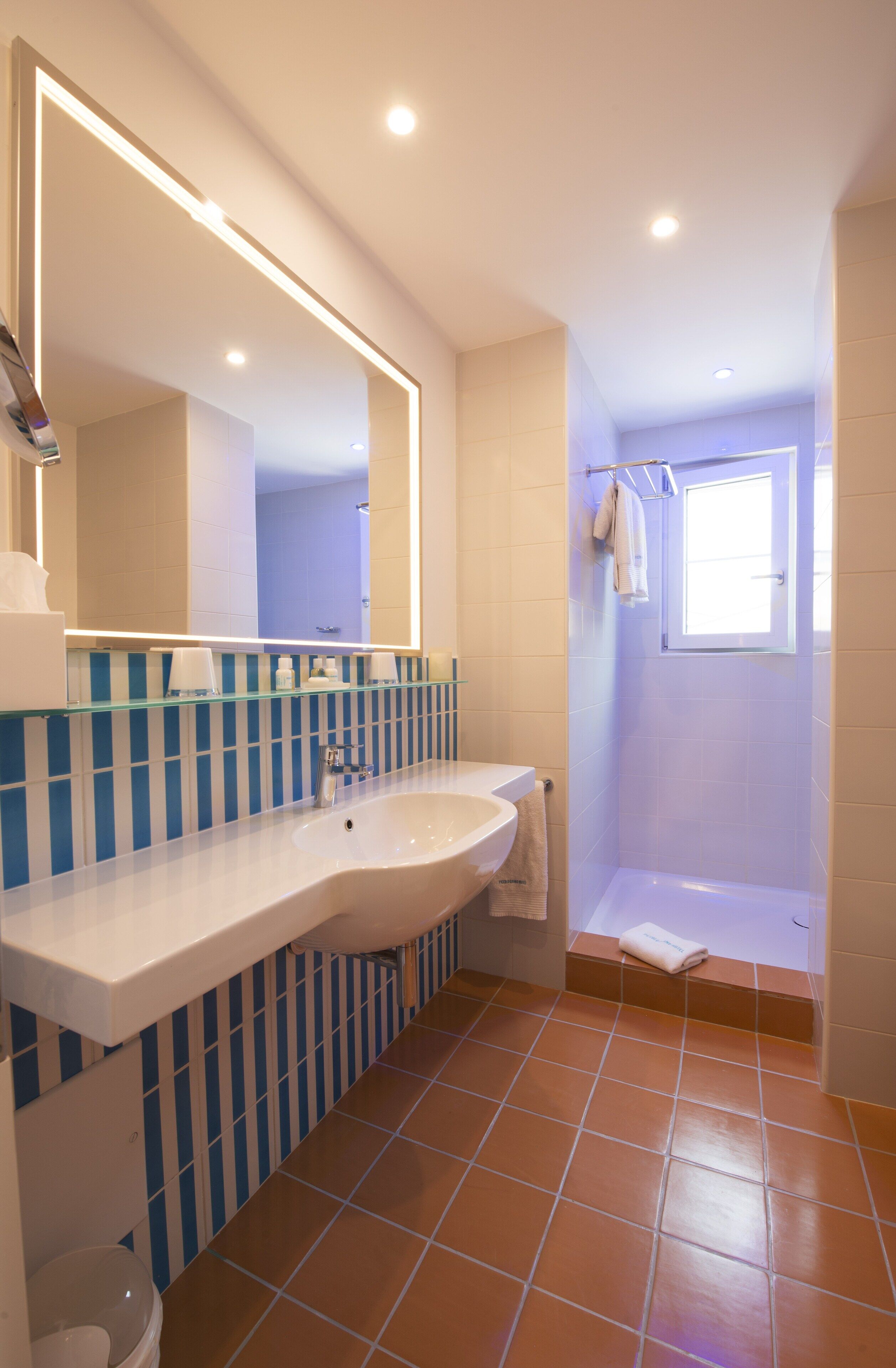 Une salle de bain avec un grand miroir, un lavabo blanc, des carreaux bleus à rayures et une fenêtre.
