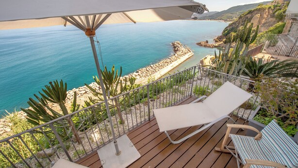 Terraza de madera con dos sillones de descanso y una mesa, con vista a la costa y una orilla rocosa con cactus.