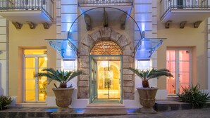 Una imponente entrada de hotel con un arco de piedra, barandillas de cristal y plantas en macetas.