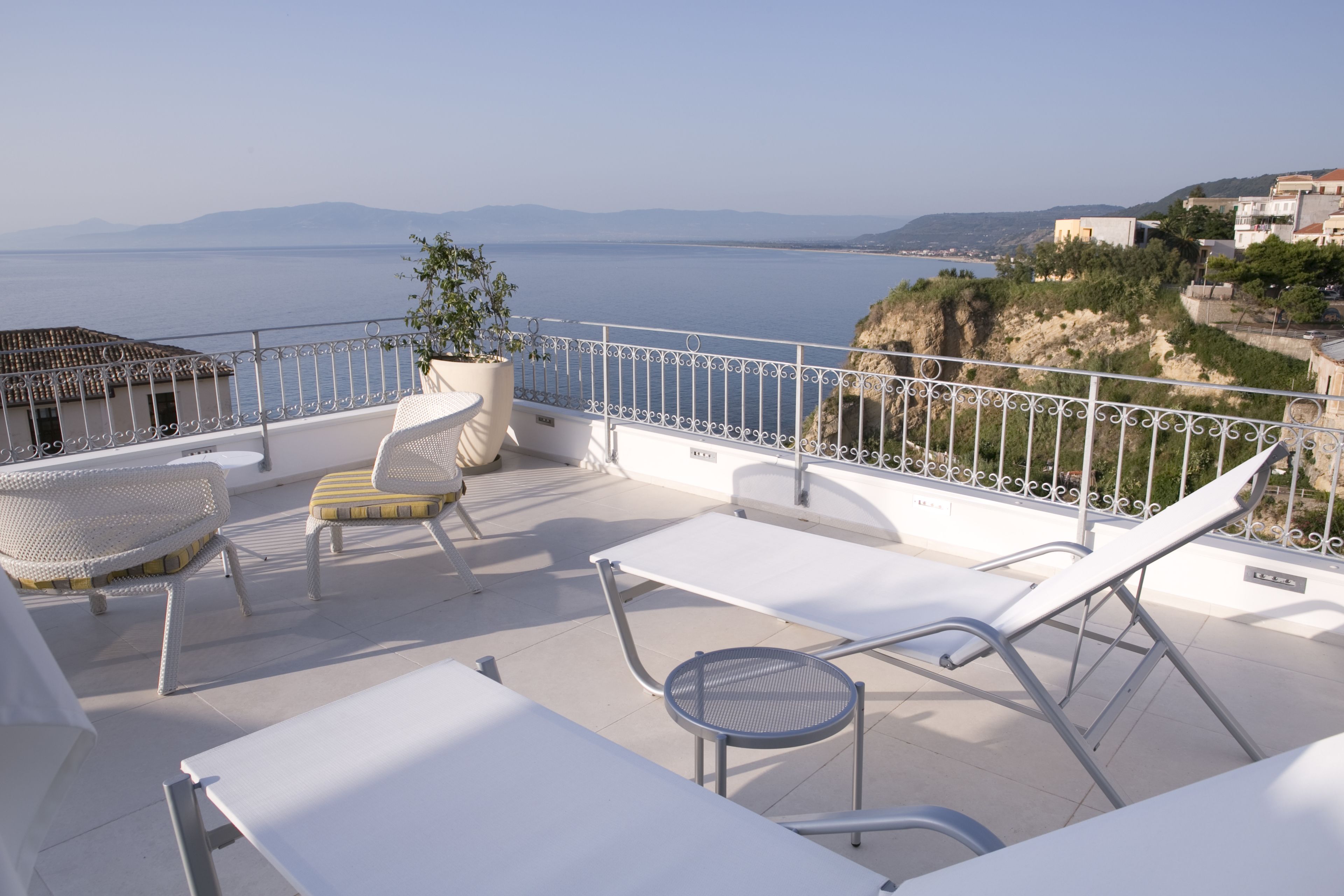 A terrace with white furniture, a potted plant, and a view of the sea and distant mountains.