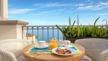 Una mesa redonda de madera con alimentos para el desayuno, dos sillas blancas de mimbre y vistas al mar.