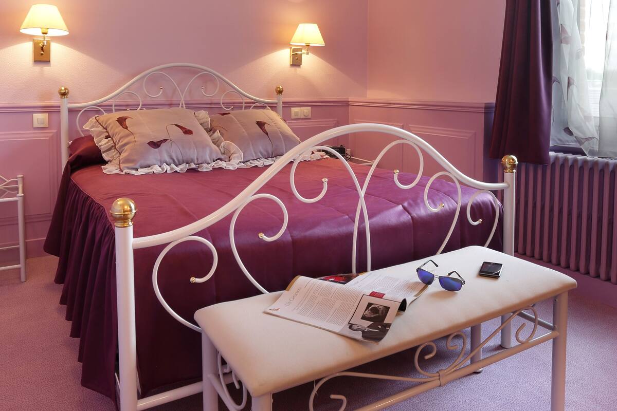 A bedroom with a bed, bedside lamps, a nightstand, a table with glasses and a book, and a window with curtains.