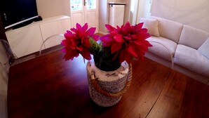Un salón con suelo de madera, un sofá blanco, un televisor sobre un mueble y un jarrón con flores rojas.