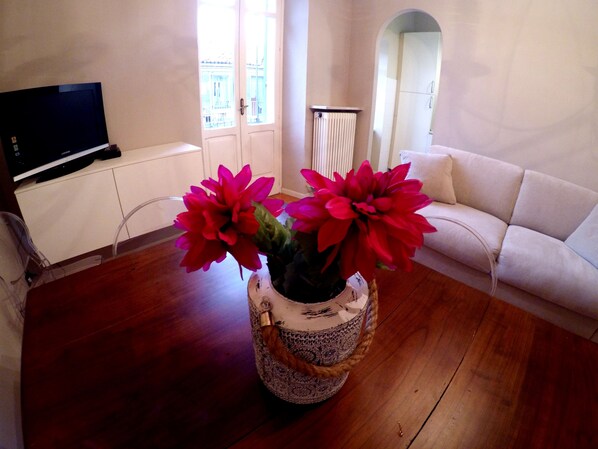 A living room with a wooden floor, a white sofa, a TV on a cabinet, and a vase with red flowers.