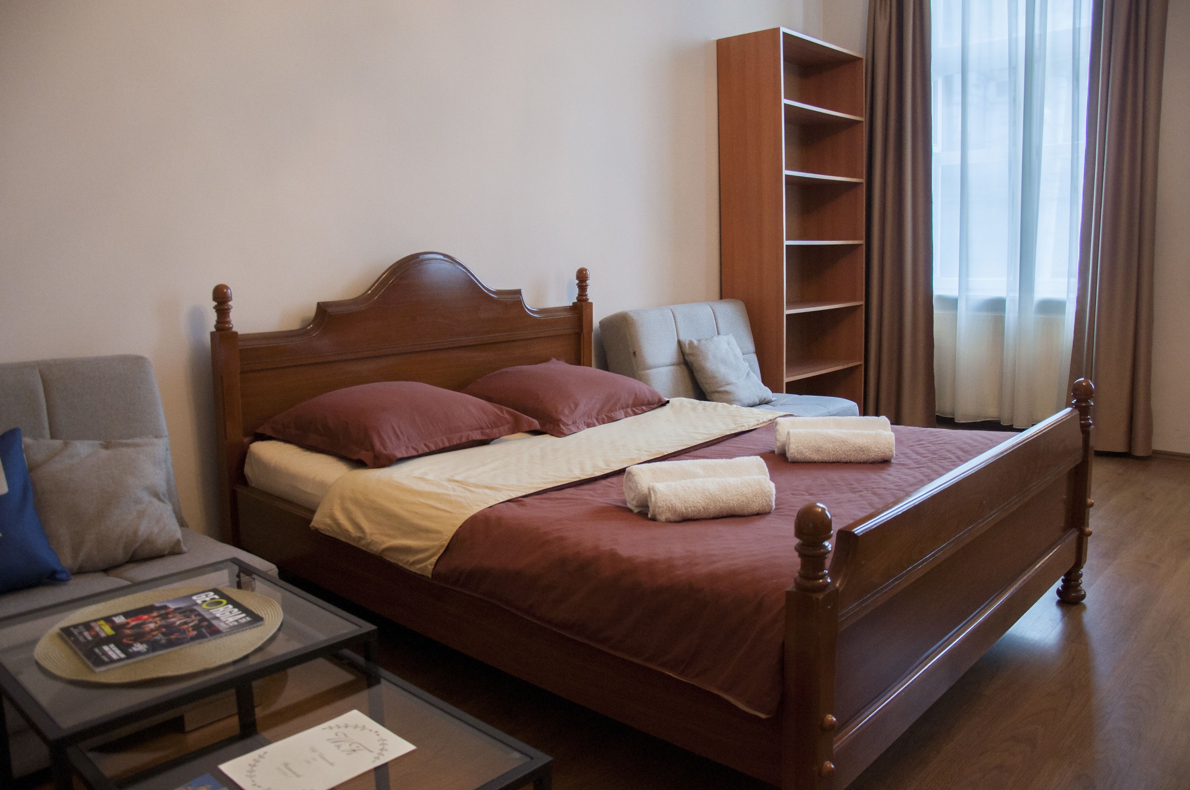 A neatly made bed with a wooden headboard, a gray armchair, a small table with a magazine, and a bookshelf.
