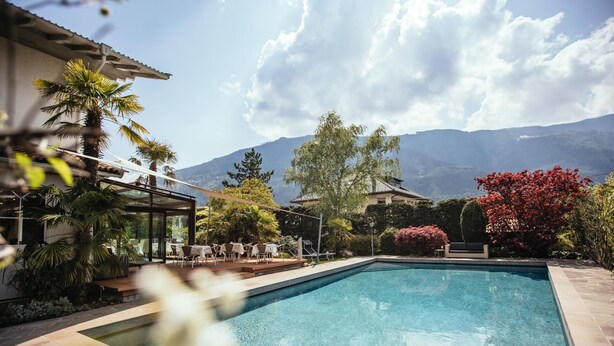 A swimming pool with a patio area, surrounded by palm trees and a mountain view. - Hotel Rablanderhof (Parcines)