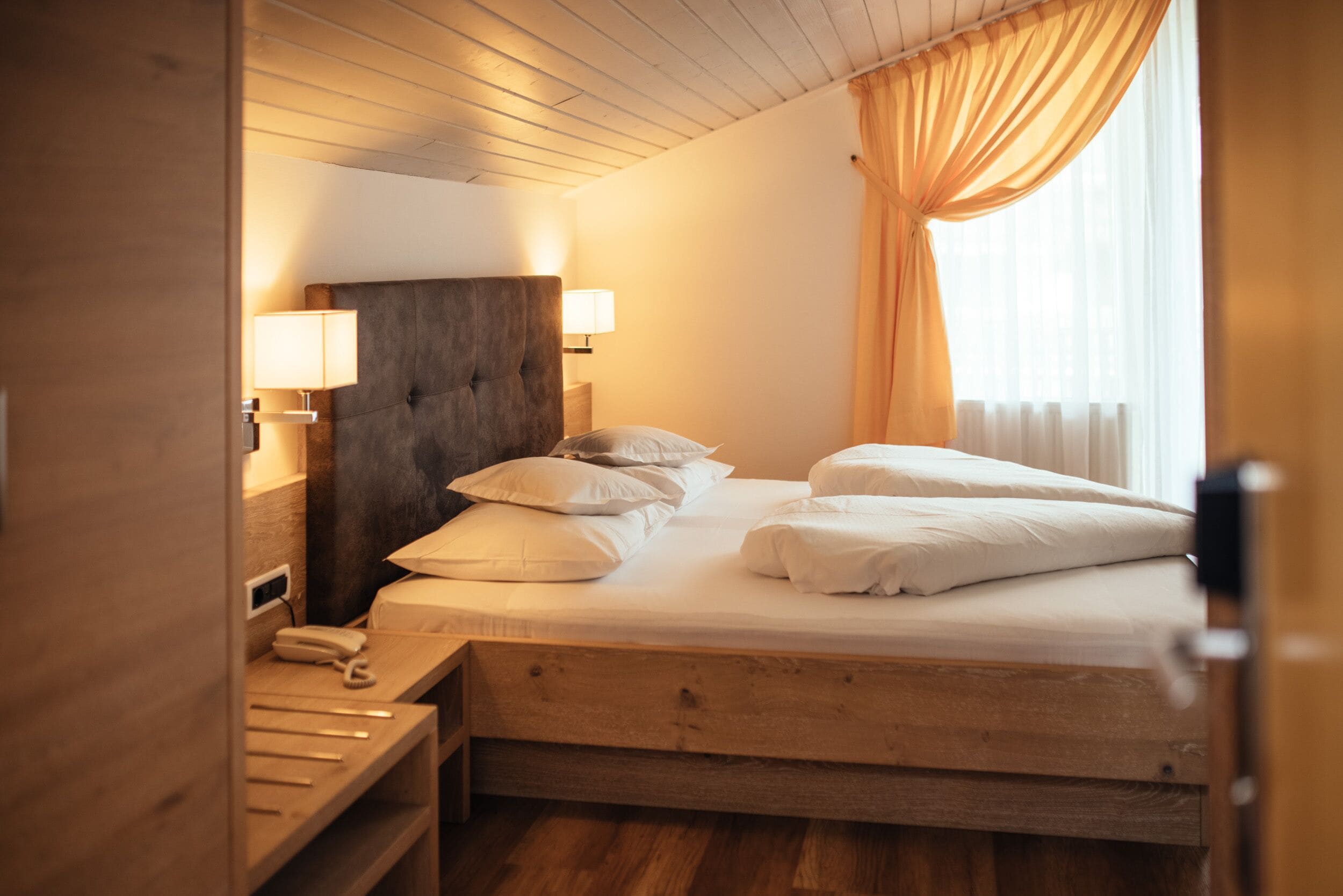 A neatly made bed with white linens and pillows, a wooden headboard, bedside lamps, and a wooden nightstand with a telephone.