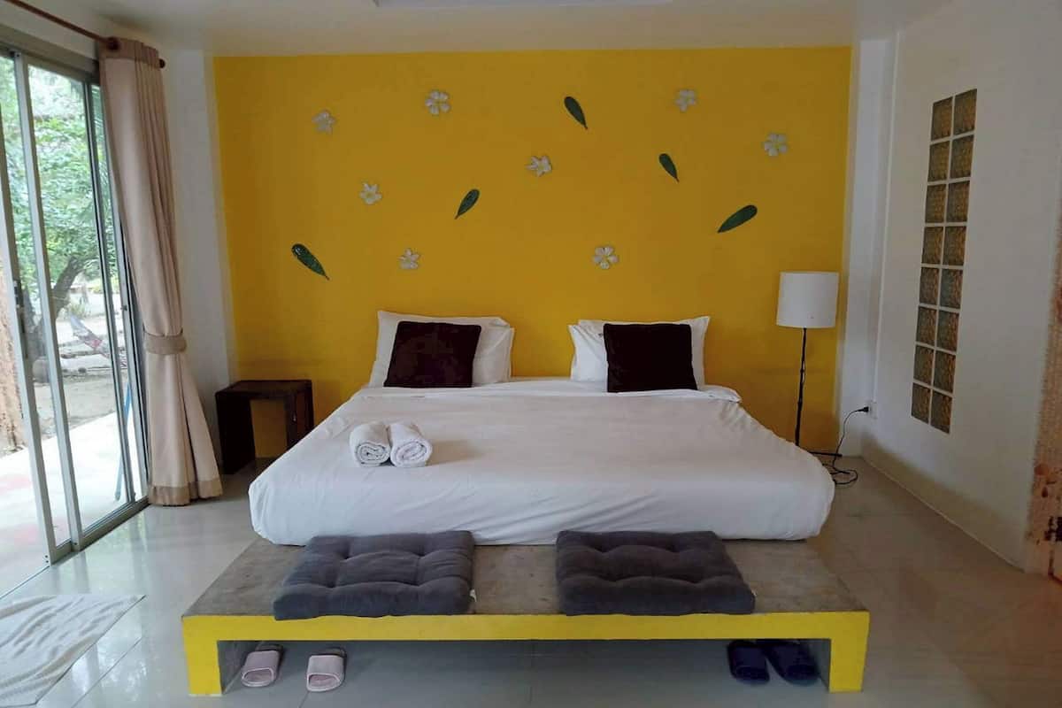 A bedroom with a bed, pillows, a bedside lamp, a small table, and a bench with towels.