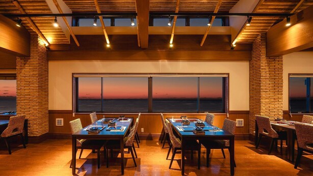 A conference room with a large window overlooking a sunset, wooden beams on the ceiling, and a brick wall. - Izu Inatori Resort Sazana (Higashiizu)