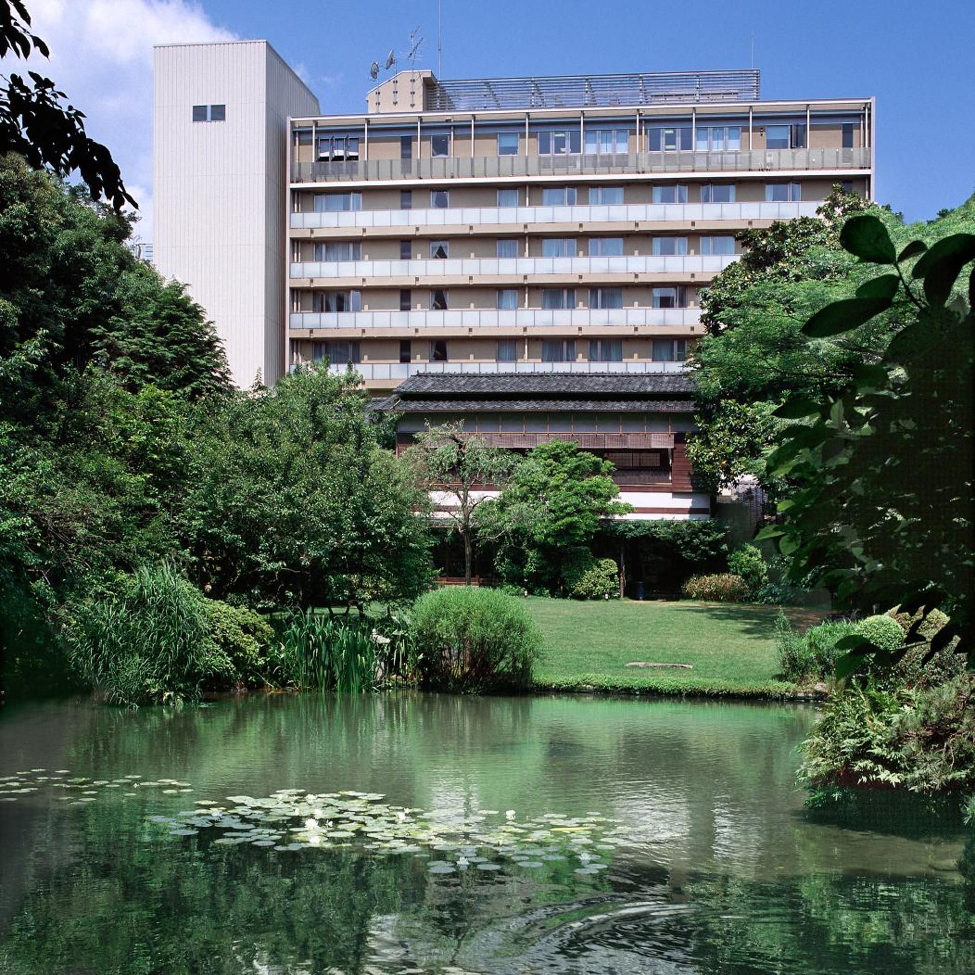 A multi-story hotel with a flat roof and balconies, surrounded by lush greenery and a pond with lily pads.