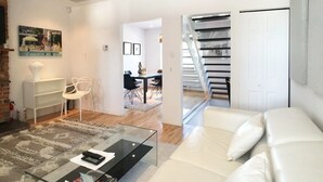 A modern living room with a glass coffee table, white sofa, and a staircase leading to an upper floor. - HPG Griffintown (Montreal)