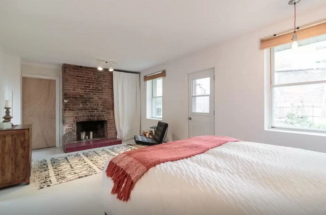 A bedroom with a brick fireplace, a large bed with a pink throw, a wooden dresser, and a view of the street through a window.