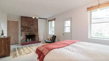 A bedroom with a brick fireplace, a large bed with a pink throw, a wooden dresser, and a view of the street through a window.