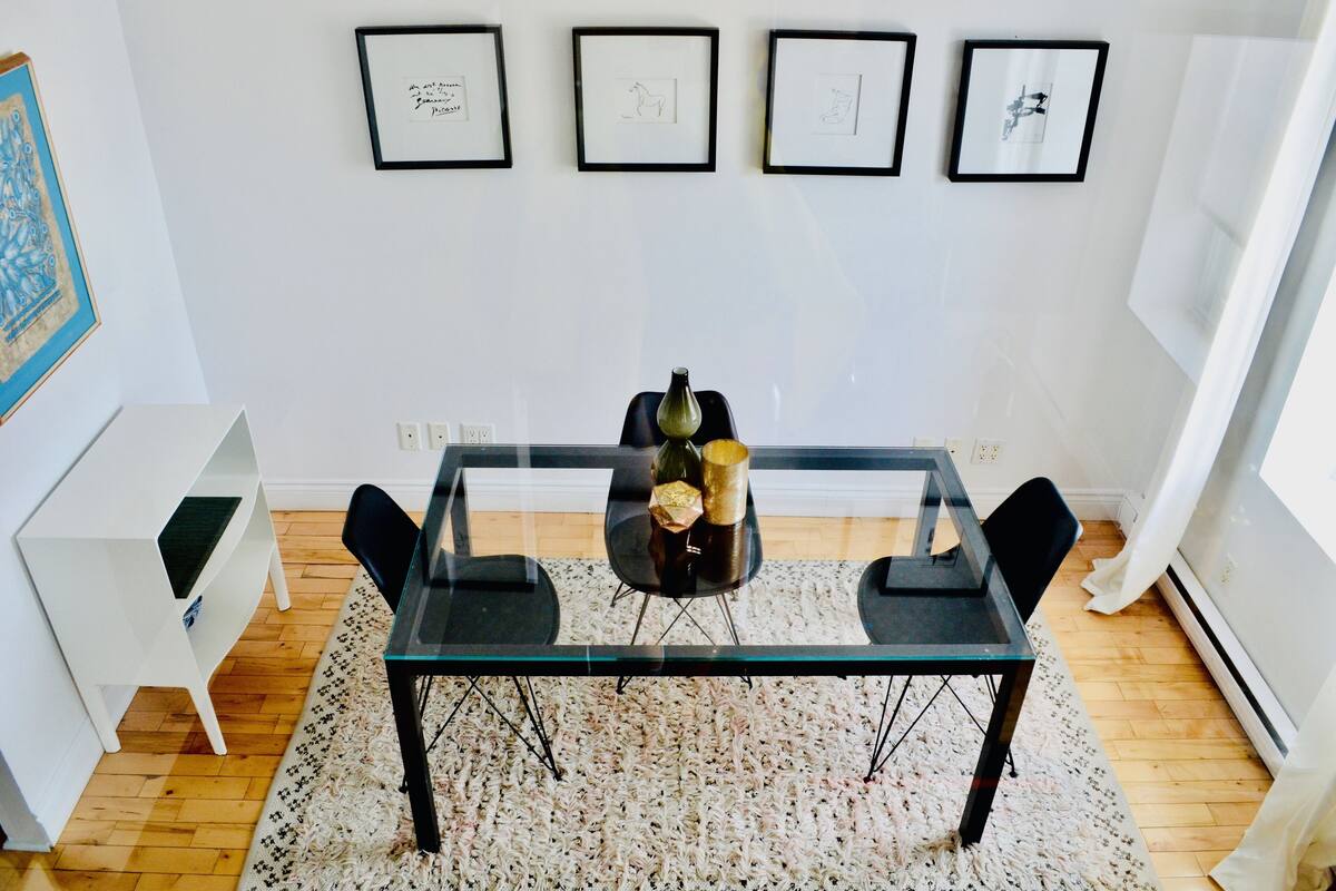 A modern dining area with a glass table, black chairs, and a white cabinet. There are framed artworks on the wall and a decorative vase on the table.