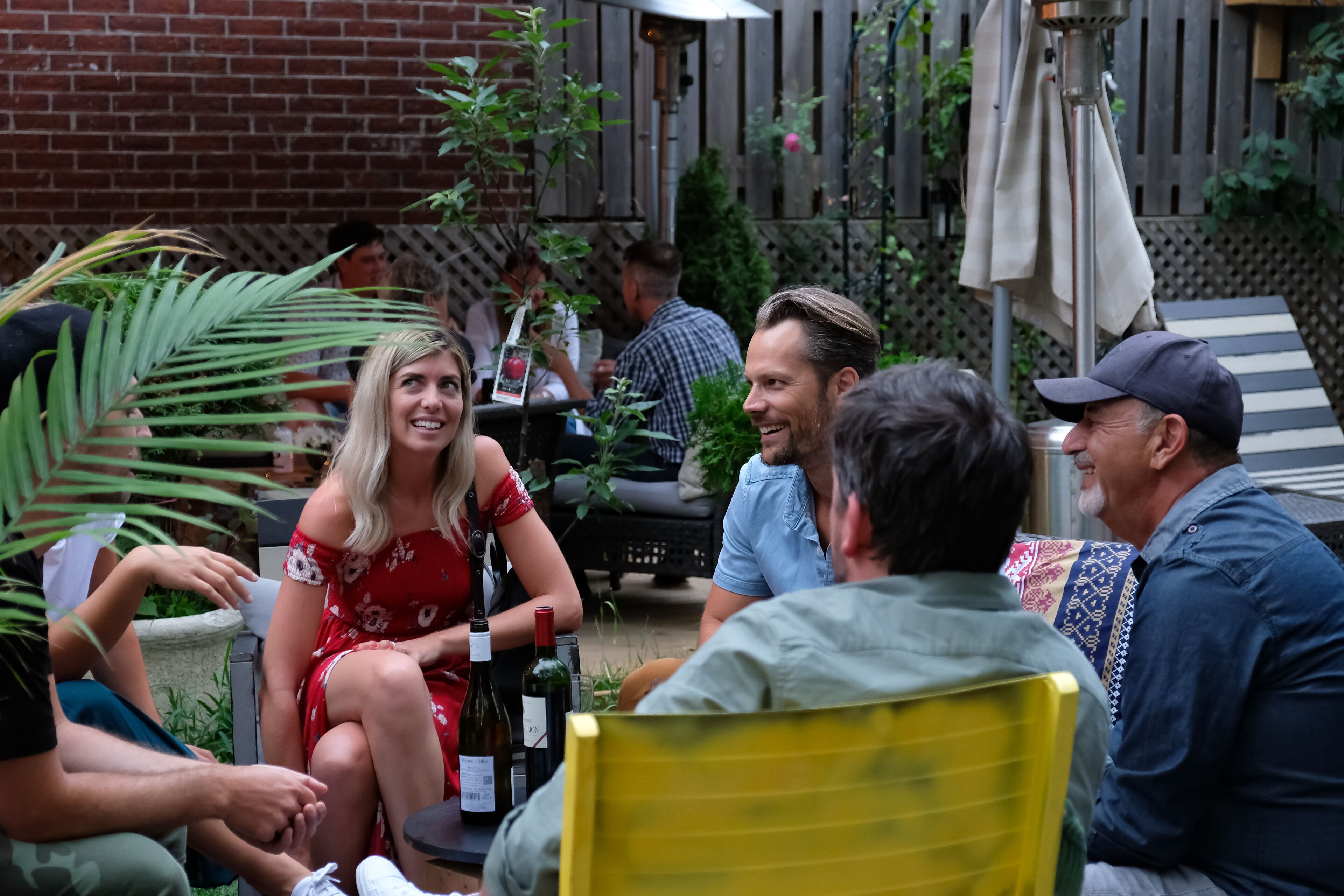 Un groupe de personnes est assis à une table à l’extérieur, autour de boissons, et est entouré de verdure et d’un mur de briques.