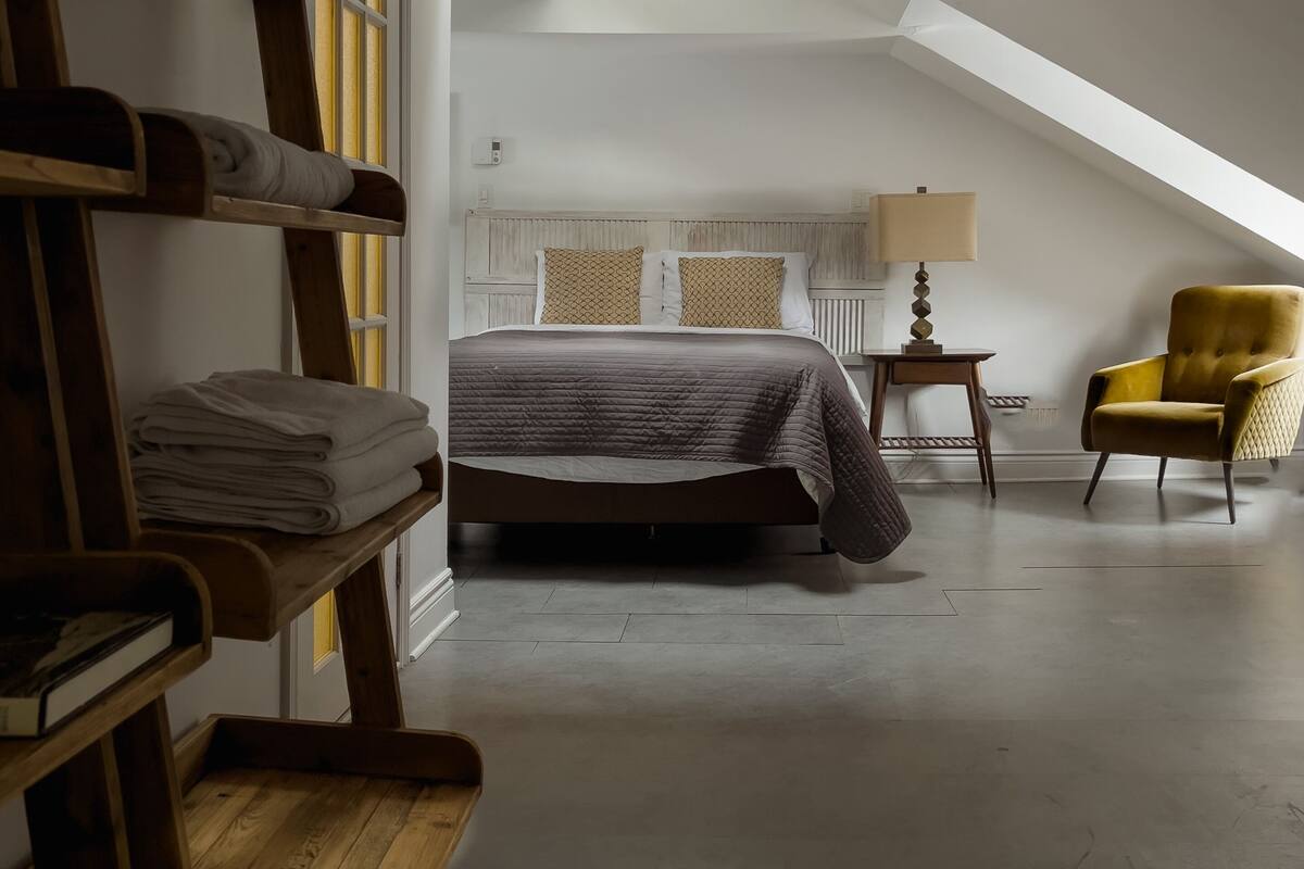 A bedroom with a bed, a chair, a lamp, and a shelf with towels.